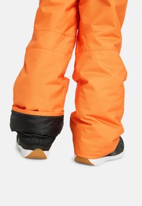 Felle oranje sneeuwbroek met een zwarte elastische manchet aan de zoom, met een gladde, waterdichte textuur en een losse pasvorm boven zwarte sportschoenen.