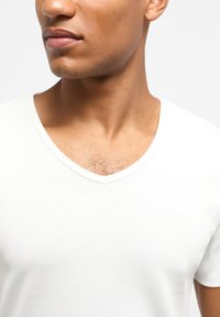 Tricou alb din bumbac cu guler în V, cu o textură netedă. Prim-planul prezintă un logo subtil pe partea stângă a pieptului, expunând o mică porțiune de păr de piept.