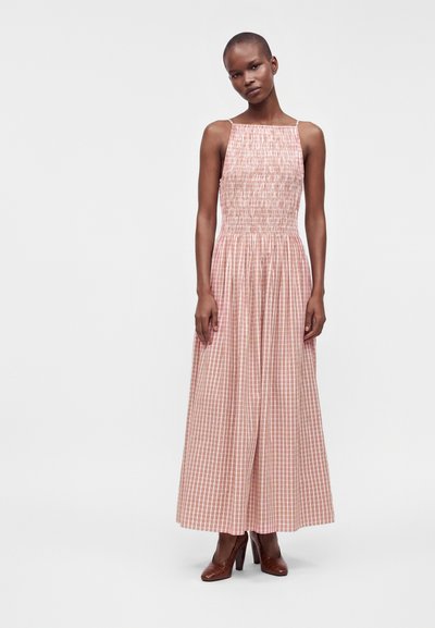 Woman wearing a sleeveless pink and white checkered maxi dress with smocked bodice and brown high-heeled shoes standing against a plain background.