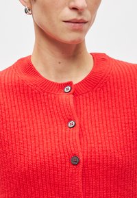 Κόκκινο ριμπε κάρντιγκαν με στρογγυλή λαιμόκοψη και τρία μαύρα κουμπιά. Η υφή είναι πλεκτή, προσφέροντας μια ζεστή και άνετη εμφάνιση.