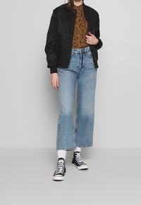 Schwarze Bomberjacke über einem braunen Leopardenmuster-Oberteil, kombiniert mit hellblauen, weit geschnittenen Jeans und schwarzen niedrigen Sneakern mit weißen Schnürsenkeln.