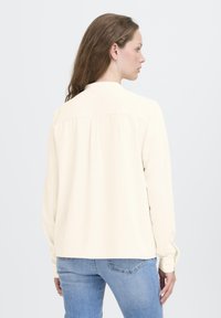 Kvinde med langt brunt hår iført en cremefarvet langærmet bluse og blå jeans, set bagfra mod en ensfarvet lys baggrund.