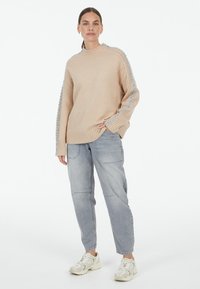 Beige knitted sweater with ribbed neckline and textured blue accents on sleeves, paired with light gray cargo pants and white sneakers.