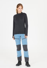 Zwarte ritsjacket van textuurstof, gecombineerd met blauwe broek met zwarte accenten en ritszakken. Zwarte veterschoenen maken de outfit compleet.