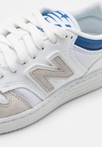 Vit sneakers med blå detaljer och en grå mocka 'N'-logotyp. Har strukturerade mesh-paneler, platt snörning och en tjock gummisula.