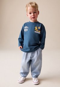 Zils krekls ar garām piedurknēm un "PAW PATROL" logo, kā arī multfilmu tēli, kopā ar vaļīgām gaiši zilām džinsa biksēm un baltām sporta kurpēm.