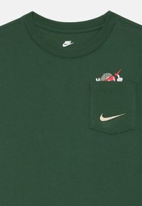 Žalias medvilninis marškinėliai su apvalia iškirpte, turintys kišenę ir spalvingą grafiką viduje. Virš kišenės yra mažas auksinės spalvos Nike logotipas.