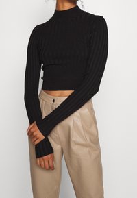 Haut court côtelé noir avec un col montant et des manches longues, arborant une coupe ajustée et un tissu texturé. Associé à un pantalon beige taille haute.