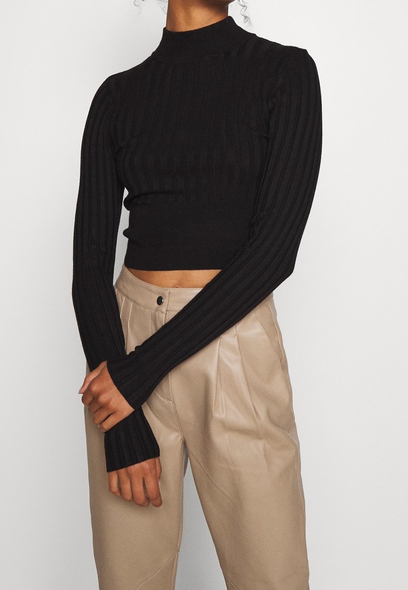 Haut court côtelé noir avec un col montant et des manches longues, arborant une coupe ajustée et un tissu texturé. Associé à un pantalon beige taille haute.