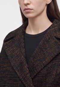 Femme portant un manteau en laine texturé marron foncé et noir par-dessus un haut noir, visible des épaules au menton sur un fond clair uni.