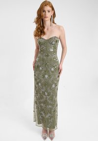 Abito da sera senza spalline di colore verde oliva con intricati ricami floreali, lavorazione di perline e silhouette aderente, abbinato a scarpe con tacco trasparente.