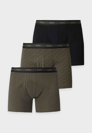 Homme à la peau foncée en forme, debout pieds nus, portant un boxer vert à motifs avec une ceinture noire sur un fond blanc uni.