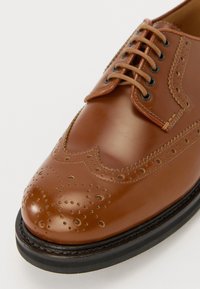Brązowe skórzane buty formalne z designem typu wingtip, z detalami brogu oraz okrągłym noskiem. Posiada sześć dziurek na sznurowadła oraz płaską czarną gumową podeszwę.