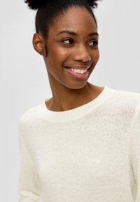 Leichter Strickpullover in Offweiß. Mit weitem, geripptem Ausschnitt und strukturiertem Stoff. Das Modell lächelt und präsentiert das Design.