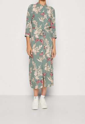Femme portant une robe chemise midi verte avec des imprimés floraux roses et des papillons, taille cintrée, manches trois-quarts, chaussettes blanches et baskets blanches.