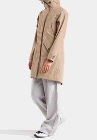 Beige vedenpitävä takki hupulla, vetoketjulla ja sivutaskuilla. Päällä löysät, vaaleanharmaat housut ja mustavalkoiset tennarit.