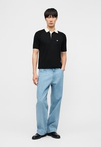 Vyras dėvi juodą trumpomis rankovėmis polo marškinėlį su baltu apykakle, šviesiai mėlynas plačias kelnes ir juodus batus, stovi su viena ranka kišenėje.