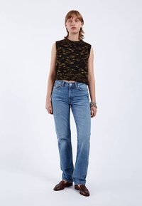 Černý a žlutý tílko s fleky v kombinaci s světle modrými džíny se širšími nohavicemi. Modelka stojí před bílým jednoduchým pozadím.