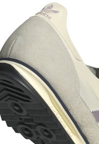 Light grey suede sneaker with cream accents and a black textured rubber sole. Features purple branding on the upper. Classic design elements.