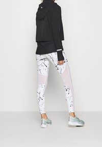 Persona che indossa un top a maniche lunghe nero e leggings bianchi con motivi astratti neri e pannelli in rete, in piedi di fronte a uno sfondo bianco uniforme.