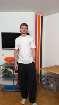 Jeune homme tatoué portant un t-shirt blanc et un pantalon noir, debout dans une pièce avec des rayures verticales colorées, une télévision et une lampe orange.