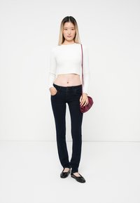 Joven mujer que lleva una camiseta blanca de manga larga corta, jeans oscuros ajustados, zapatos planos negros y sostiene un pequeño bolso de hombro burdeos, de pie contra un fondo blanco.