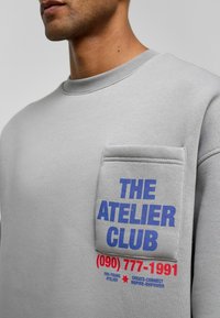 Grijze sweatshirt met een ronde hals, een voorkantzak en gedurfde tekstgrafieken in blauw en rood. Katoenen materiaal met een soepele textuur.