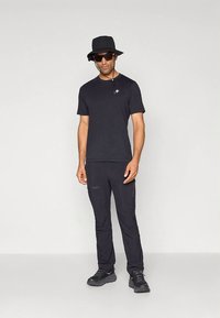 Černé tričko s krátkým rukávem ve spojení s černými cargo kalhotami, černým kloboukem typu bucket a tmavými slunečními brýlemi. Boty mají strukturovaný povrch.