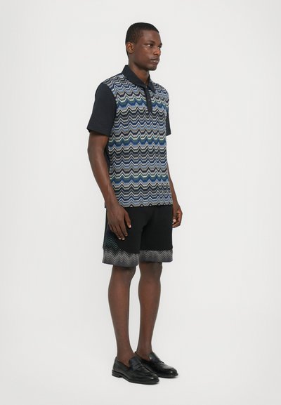 Cămașă polo cu mânecă scurtă, cu un model în zigzag în nuanțe de albastru și verde, combinată cu pantaloni scurți negri ce au un tiv în zigzag asortat, purtată cu loafers negri.
