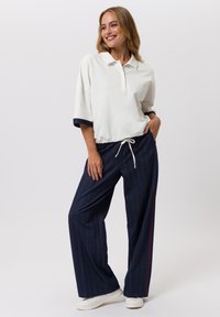 Chemise polo blanche à coupe décontractée avec des manches retroussées, associée à un pantalon large à rayures sombres avec une accent rouge sur le côté. Baskets blanches.