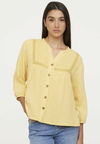 Blouse jaune à boutons avec un design texturé et plissé, ornée de dentelle, présentant des manches trois-quarts et une coupe décontractée.