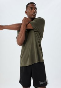 Olijfgroene shirt met korte mouwen en een losse pasvorm, gecombineerd met zwarte shorts met een kleine tekstafdruk en grijze zijpanelen.
