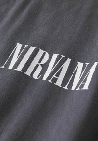Zwarte katoenen stof met grote, opvallende witte tekst met de tekst "NIRVANA" in een gestileerd schreeflettertype. De tekst is gecentreerd en licht versleten.