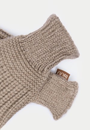Große beige gestrickte fingerlose Handschuhe mit gerippten Bündchen und einem sichtbaren braunen Stoffetikett auf weißem Hintergrund.