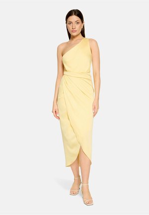 Kobieta ubrana w bezrękawową, asymetryczną, bladożółtą sukienkę midi z marszczonym talią i tulipanowym dołem, zestawioną z beżowymi szpilkami z paskiem wokół kostki.