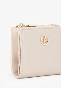 Portafoglio in pelle beige con finitura testurizzata, accessori in oro e un dettaglio logo circolare. Chiusura a cerniera lungo la parte superiore. Forma compatta e rettangolare.