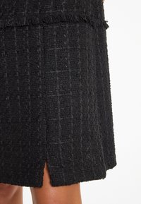 Schwarzer Tweedrock mit strukturiertem Oberflächen, der ein dezentes klassisches Muster und Fransenverzierungen am Saum aufweist, und mit einem Schlitz für mehr Bewegungsfreiheit.