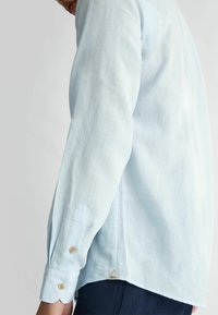 Camicia di lino azzurro chiaro con maniche lunghe, polsini abbottonati e orlo curvo. Presenta un piccolo etichetta logo sul lato.