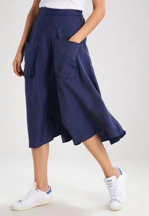 Person wearing a flowing navy blue striped skirt with large front pockets and white sneakers walking against a plain background.