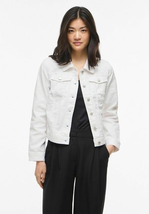 Jauna moteris, turinti vidutinio ilgio tamsius plaukus, dėvi baltą džinsinę striukę su sagomis ant juodo džemperio ir juodas plačių kojų kelnes, stovėdama su ranka kišenėje.