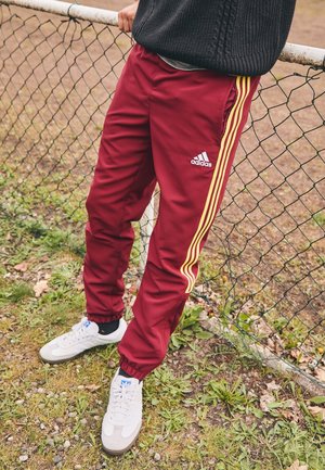 Osoba w bordowych spodniach dresowych Adidas z żółtymi paskami i białych sneakersach stoi przy siatce na trawie i ziemi.