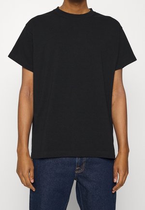 Persoon die een effen zwart T-shirt met korte mouwen en een blauwe spijkerbroek draagt, staand tegen een witte achtergrond met de armen ontspannen langs het lichaam.