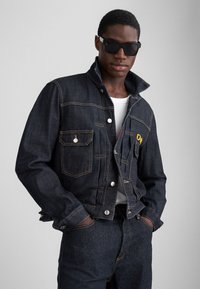 Chaqueta de mezclilla oscura con un cuello alto, cierre frontal con botones y dos bolsillos en el pecho. Presenta detalles bordados en amarillo y costuras en contraste.