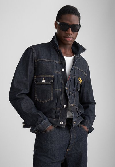Veste en denim foncé avec un col haut, un devant à boutons et deux poches poitrine. Présente des détails brodés jaunes et des coutures de contraste.
