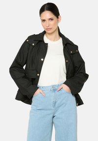 Juodas nepralaidus vandeniui švarkas su apykakle, auksinės spalvos detalių akcentais ir dviem priekinėmis kišenėmis, derinamas su baltu marškinėliu ir mėlynomis džinsomis.