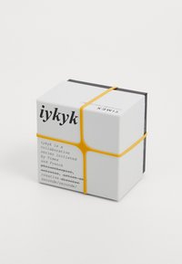 Boîte cubique blanche avec un élastique jaune, comportant un texte noir détaillant une série collaborative de Timex. La boîte a une finition matte.