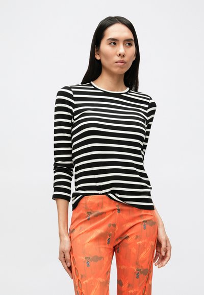 Melns un balts svītrains krekls ar garām piedurknēm un apaļu izgriezumu, kopā ar oranžām rakstainām biksēm ar dekoratīvām detaļām.