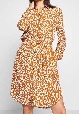 Frau trägt ein orange-weißes, leopardenmusterndes Hemdkleid mit langen Ärmeln und Taillengürtel, steht mit einer Hand in der Tasche.