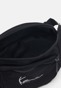 Mustapolyesterikassi, jossa on vetoketjukiinnitys, sileä pinta ja sisävuori. Etupuolella valkoinen signeerauslogo.