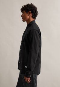 Črna majica z dolgimi rokavi iz bombaža. Ima zapenjanje spredaj s peci, bele gumbe in oznako na strani. Ljubka, tesno prilegajoča silhueta.